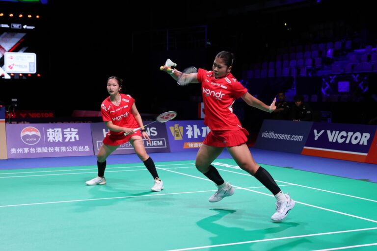 Two female badminton players in red uniforms compete on a bright green court, one reaching with her racket for a shot, shuttlecock in play.
