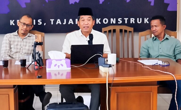 Three men sit at a wooden conference table; center man in white shirt speaks into a microphone with a laptop in front of him.
