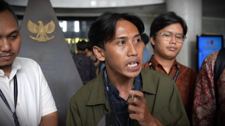 Man in a green jacket speaking, others listen in the background; a leader addressing a group in a lobby or hallway with lanyards visible.