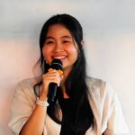 Smiling young woman with long black hair holding a microphone and speaking on stage or at a presentation.