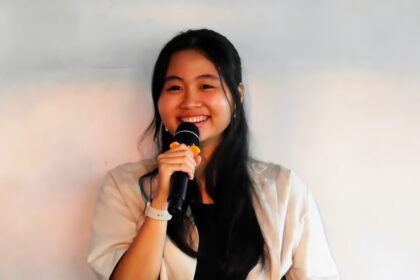 Smiling young woman with long black hair holding a microphone and speaking on stage or at a presentation.