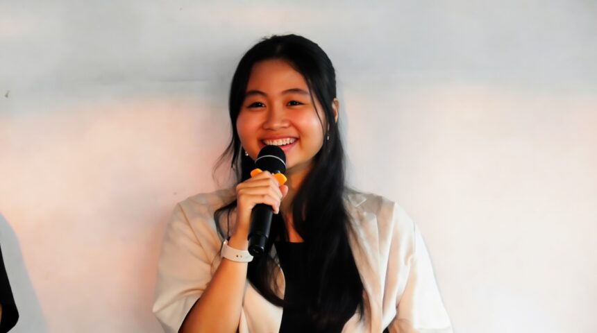 Smiling young woman with long black hair holding a microphone and speaking on stage or at a presentation.