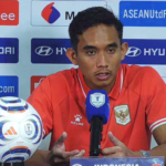Soccer player in a red jersey sits at a press conference behind a branded backdrop, speaking into a blue-microphone with a shield logo; a soccer ball rests on the table to his left.