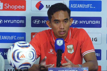 Soccer player in a red jersey sits at a press conference behind a branded backdrop, speaking into a blue-microphone with a shield logo; a soccer ball rests on the table to his left.