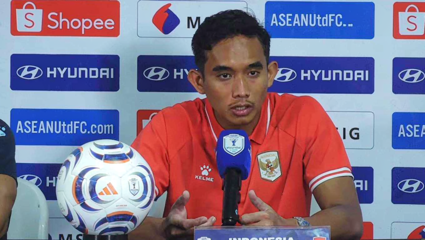 Soccer player in a red jersey sits at a press conference behind a branded backdrop, speaking into a blue-microphone with a shield logo; a soccer ball rests on the table to his left.