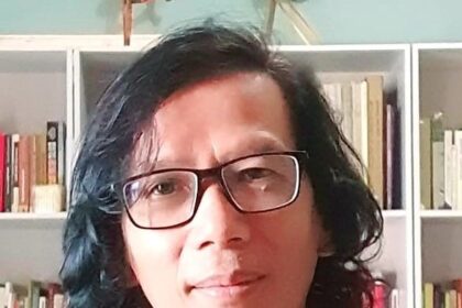 Portrait of a person with shoulder-length dark hair and glasses, standing in front of a bookshelf.