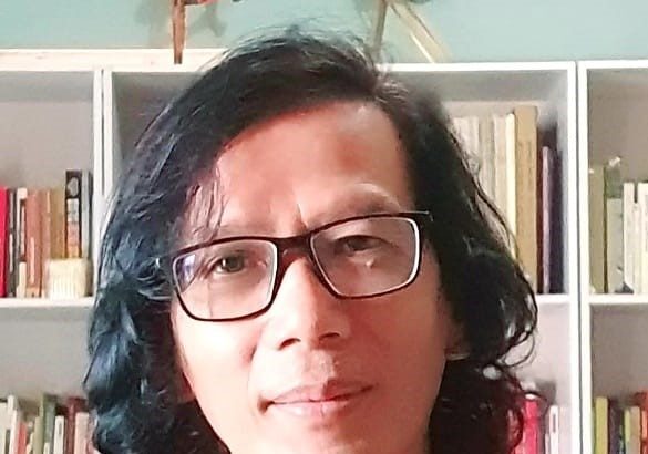 Portrait of a person with shoulder-length dark hair and glasses, standing in front of a bookshelf.