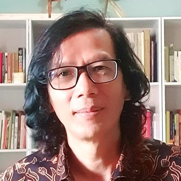Portrait of a person with shoulder-length dark hair and glasses, standing in front of a bookshelf.