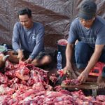 Two men chopping and sorting large chunks of raw meat on a wooden board, surrounded by a heap of red meat under a tarp backdrop.