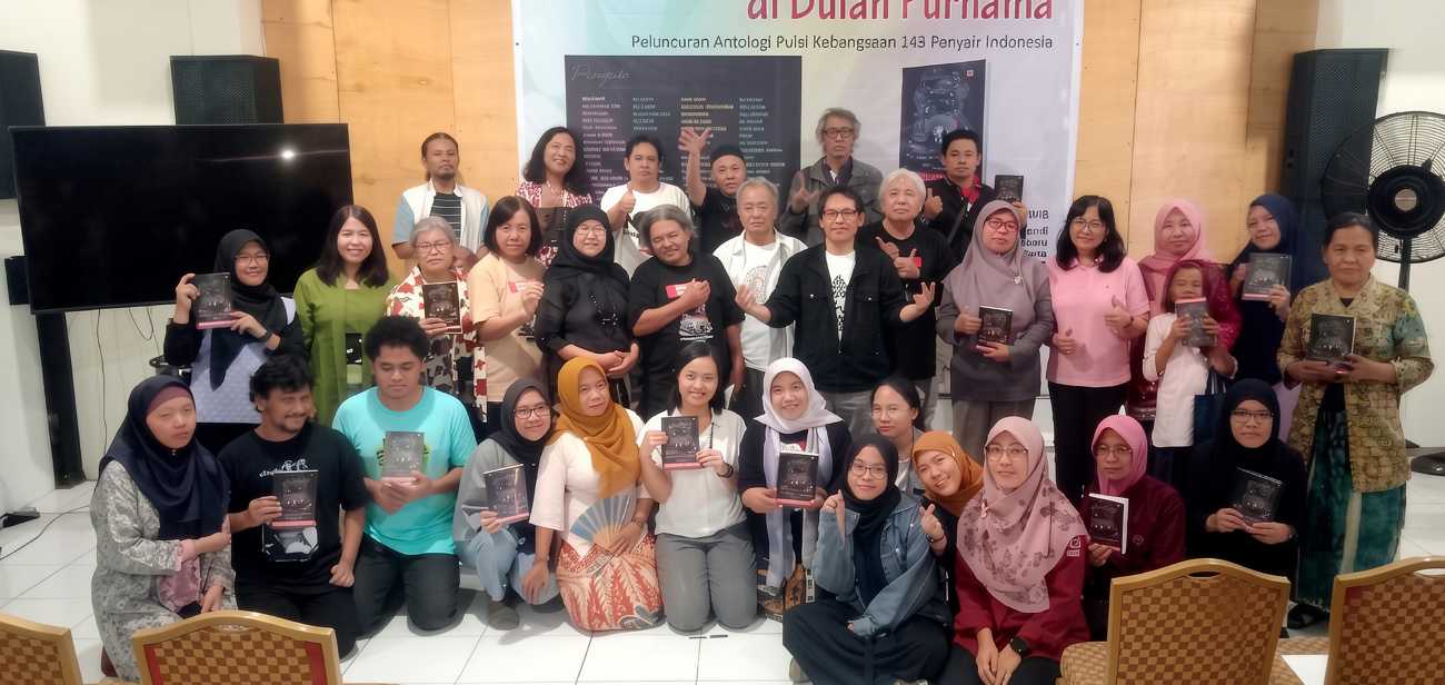 Group of about 40 people posing with books in a hall, a banner behind them indicating a national anthology event in Indonesian.