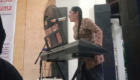Woman singing into a microphone while playing a keyboard on a stage with a stand fan nearby.