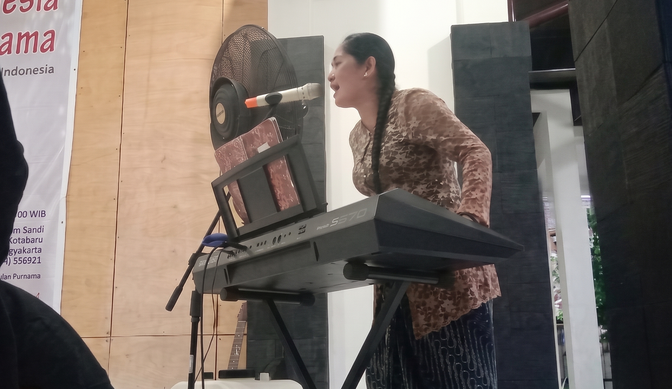 Woman singing into a microphone while playing a keyboard on a stage with a stand fan nearby.
