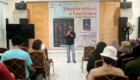 Man standing on a stage giving a talk with a large banner behind reading Manuskrip Indonesia di Bulan Purnama, audience seated in front.