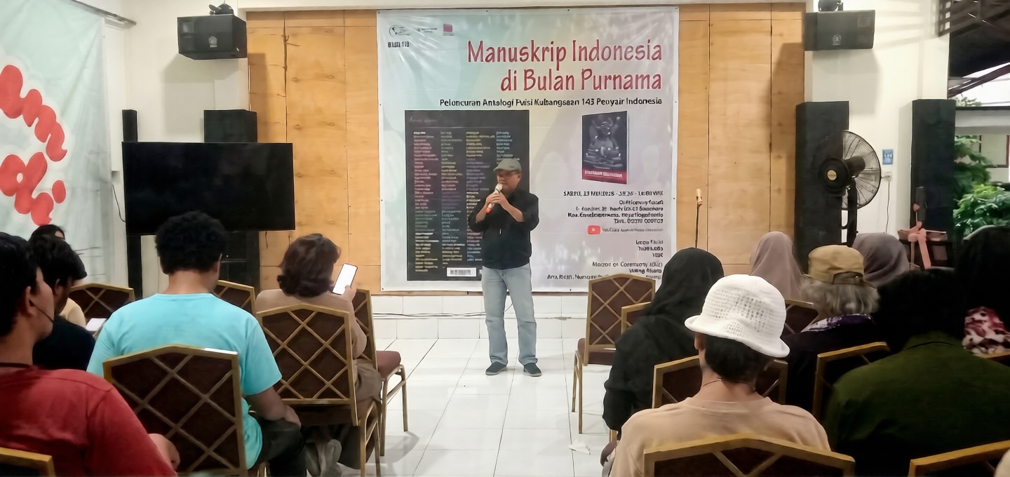 Man standing on a stage giving a talk with a large banner behind reading Manuskrip Indonesia di Bulan Purnama, audience seated in front.