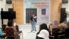Man speaking on a stage in front of a banner reading 'Manuskrip Indonesia di Bulan Purnama' with seated audience taking photos.