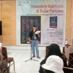 Man speaking on a stage in front of a banner reading 'Manuskrip Indonesia di Bulan Purnama' with seated audience taking photos.