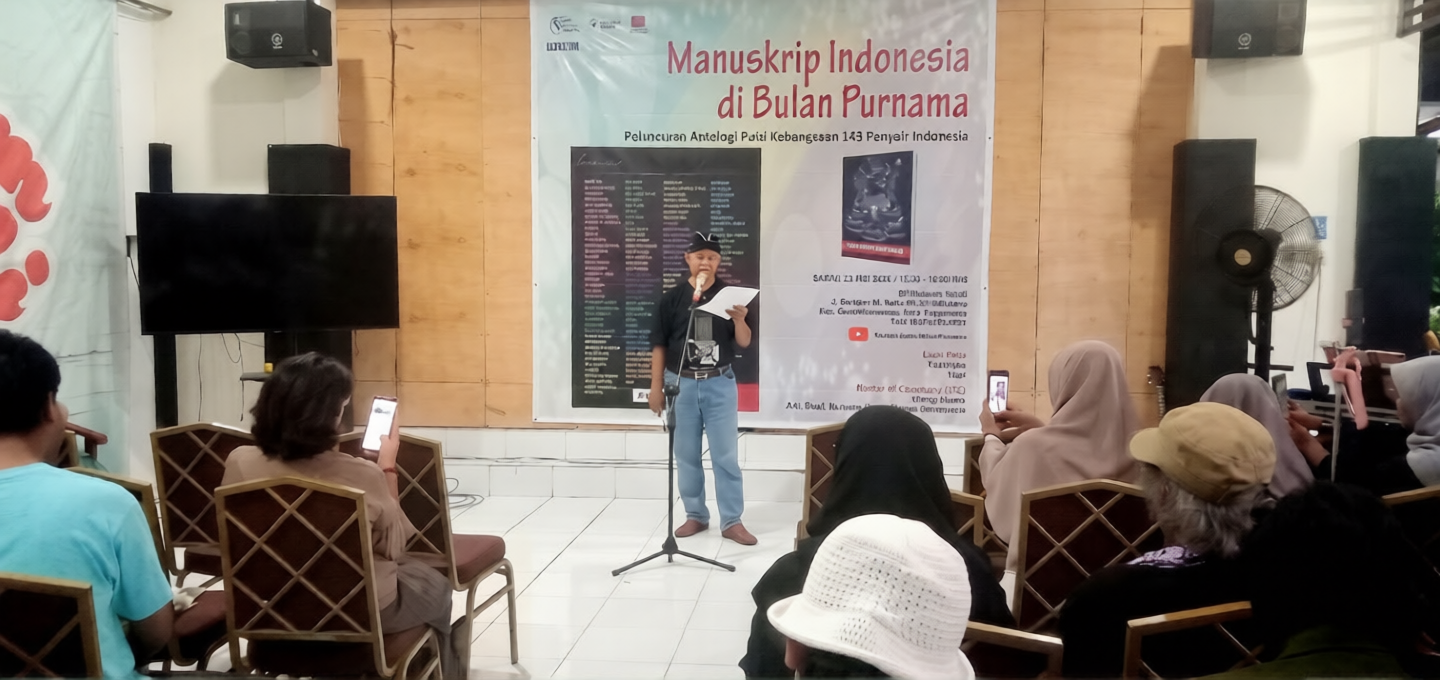 Man speaking on a stage in front of a banner reading 'Manuskrip Indonesia di Bulan Purnama' with seated audience taking photos.
