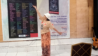 Woman dancing with one arm raised, wearing a beige T-shirt, white hat, and patterned skirt, in front of a large event banner inside a hall; a guitar rests on a stand to the right.