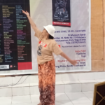Woman dancing with one arm raised, wearing a beige T-shirt, white hat, and patterned skirt, in front of a large event banner inside a hall; a guitar rests on a stand to the right.