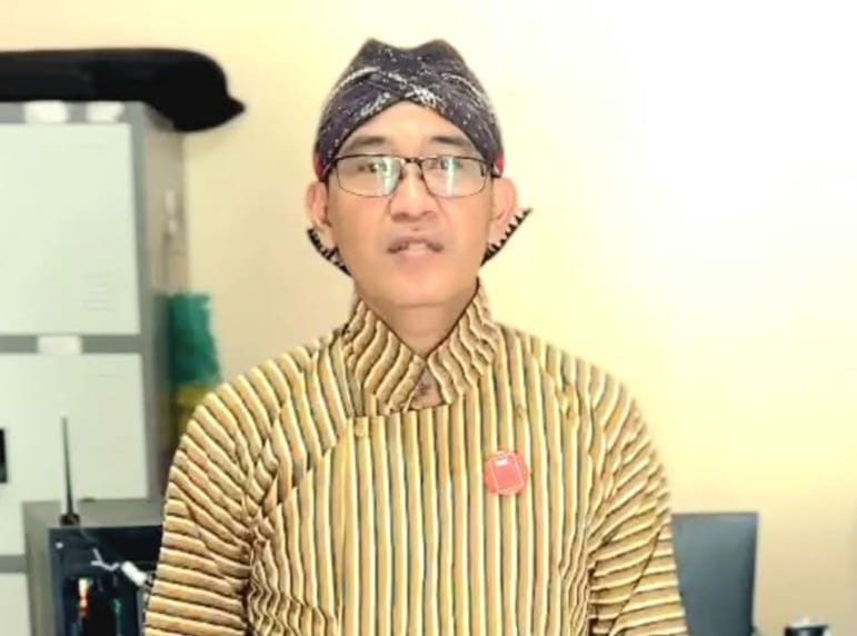 Man wearing striped traditional outfit and a headwrap, with glasses, standing in an office. A red badge is pinned to his chest.