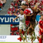 Soccer players jump to head the ball during a match with a cheering crowd in the stands behind them.