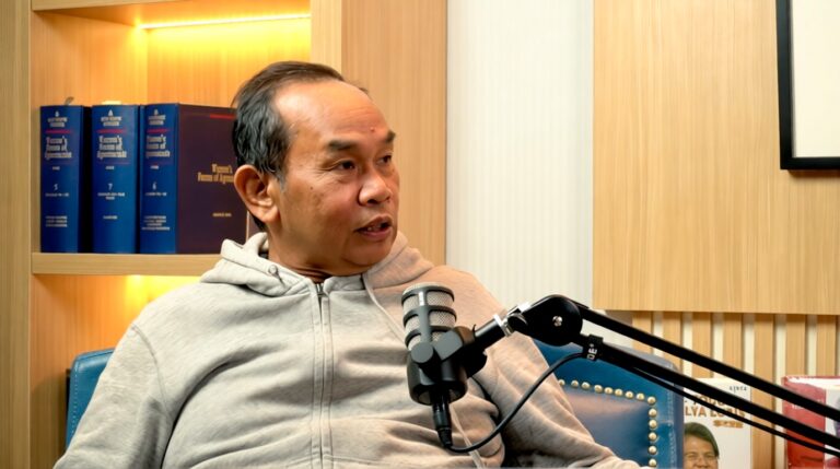 Man in a gray hoodie sits in a podcast studio, speaking into a mounted microphone with a blue chair nearby.
