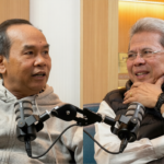 Two men in a podcast studio sit across from each other, speaking into shared microphones.