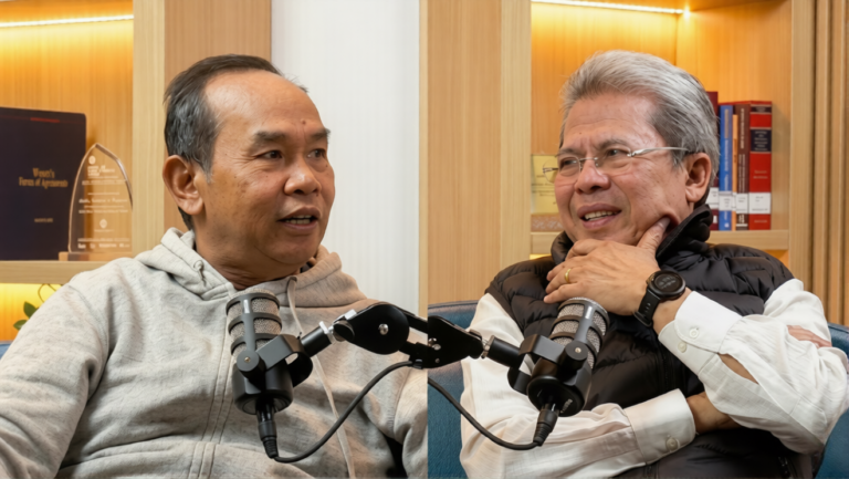 Two men in a podcast studio sit across from each other, speaking into shared microphones.
