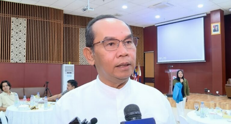 Man in a white shirt speaks to reporters in a conference room, microphones held up to him, with round tables and a projector screen in the background.