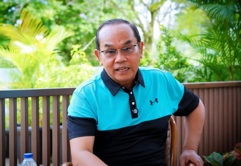 Man wearing a turquoise and black polo shirt sits on a wooden chair outdoors with lush green plants in the background, speaking or posing for a photo.