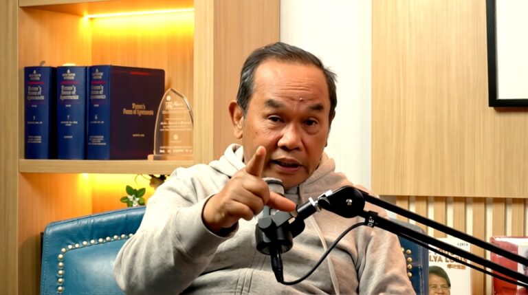 Man recording or speaking into a desk microphone in a studio setup with a blue chair and wooden shelves behind him