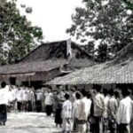 Crowd of villagers standing in a line during a flag-raising ceremony in a rural courtyard with traditional tiled-roof houses nearby.”] ,
