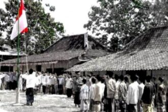 Crowd of villagers standing in a line during a flag-raising ceremony in a rural courtyard with traditional tiled-roof houses nearby.”] ,