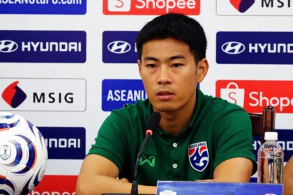 Asian male footballer in green polo speaking at a press conference with logos (Hyundai, Shopee, ASEAN) in the background and a soccer ball on the left.