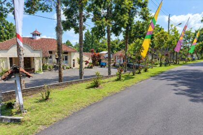 Street view of a village main road with a cream building and red-tiled roof on the left, and colorful flags along the road on a sunny day