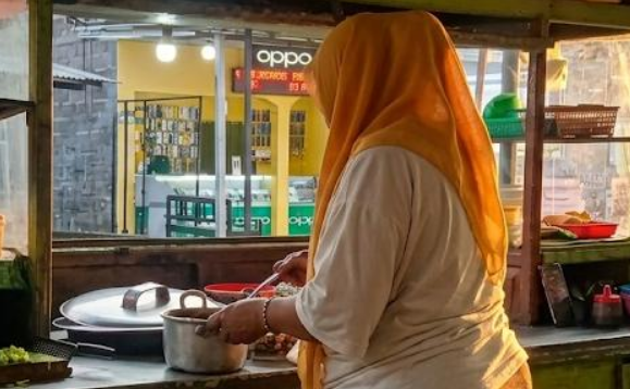 Screenshot 2026-06-12 at 00-50-20 WhatsApp-Image-2026-06-11-at-215654jpeg JPEG Image 927 × 475 pixels - Mabur.co Woman in an orange headscarf cooking at a street food stall, pot on the stove and utensils nearby.
