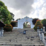 Stone stairway leads up to a white arched gate, framed by green trees and trimmed shrubs, with a sign placed on the steps.