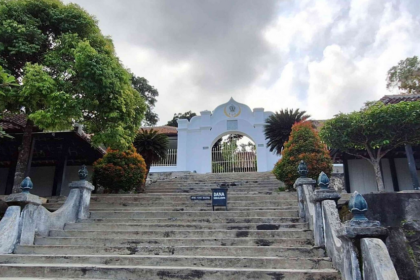 Stone stairway leads up to a white arched gate, framed by green trees and trimmed shrubs, with a sign placed on the steps.