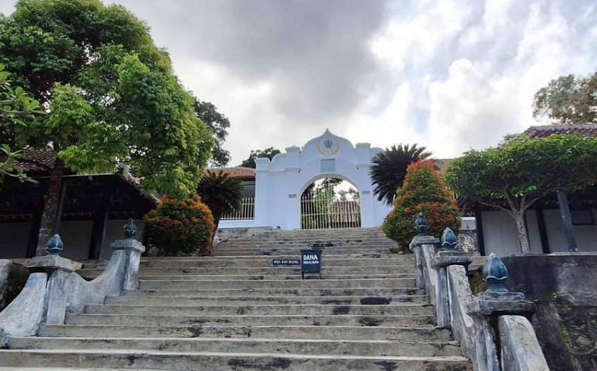 Stone stairway leads up to a white arched gate, framed by green trees and trimmed shrubs, with a sign placed on the steps.