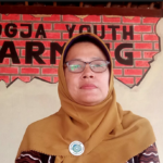Woman in a brown hijab and glasses stands before a brick wall mural that reads 'FARMING'.