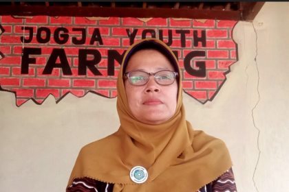 Woman in a brown hijab and glasses stands before a brick wall mural that reads 'FARMING'.