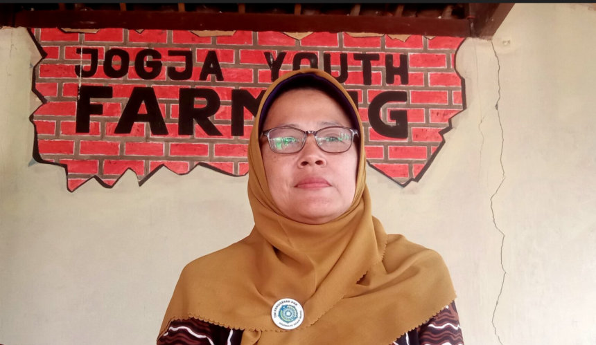 Woman in a brown hijab and glasses stands before a brick wall mural that reads 'FARMING'.