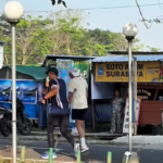 Two men jogging on a sidewalk past market stalls and a scooter; one wearing a dark sleeveless top and tights, the other in a white shirt and shorts.