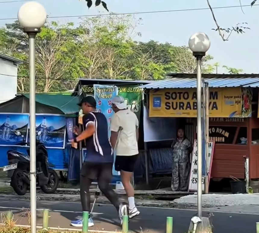 Two men jogging on a sidewalk past market stalls and a scooter; one wearing a dark sleeveless top and tights, the other in a white shirt and shorts.