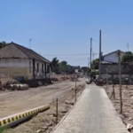 A rural residential street under construction: a narrow concrete sidewalk runs down the center with dirt and rubble on both sides.