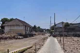 A rural residential street under construction: a narrow concrete sidewalk runs down the center with dirt and rubble on both sides.