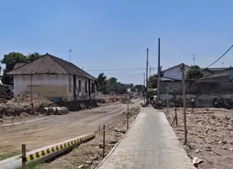 A rural residential street under construction: a narrow concrete sidewalk runs down the center with dirt and rubble on both sides.