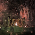Group of people walking along a water-lined path at night, illuminated by many small flames and hanging lights, creating a festival-like glow.