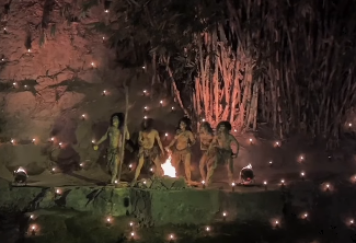 Group of people walking along a water-lined path at night, illuminated by many small flames and hanging lights, creating a festival-like glow.