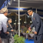 Man in a dark suit and traditional cap bows to a guest at a formal ceremony under a blue-and-white tent.